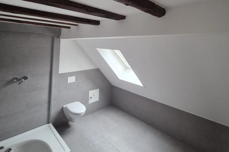 Lüftung im Bad: Dachgeschossbad mit Dachfenster, dunklen Holzbalken und grauen Wandfliesen